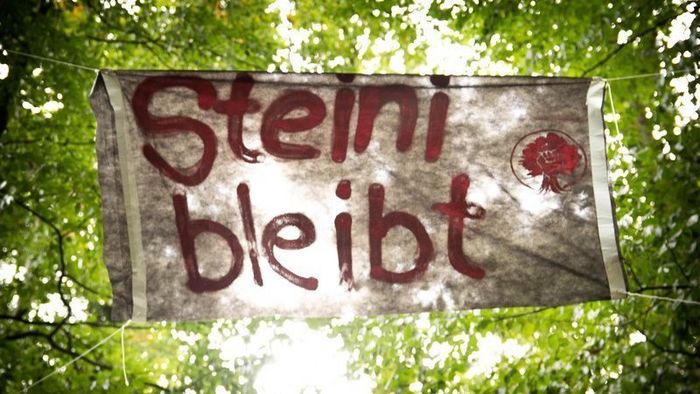 Banner vor Blick auf grüne Baumkronen mit dem Text in Rot: "Steine bleibt"